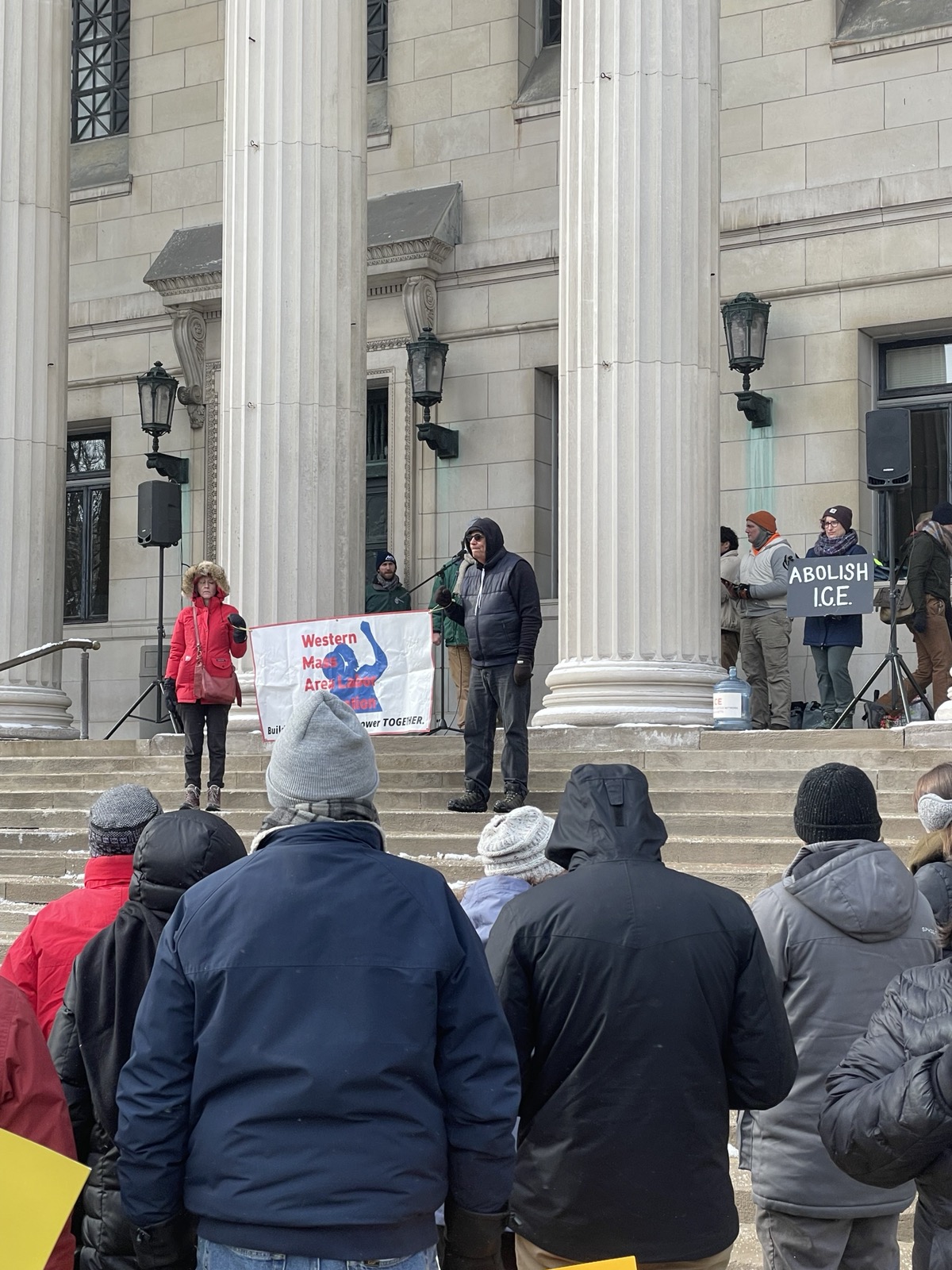 Organizers hold a Western Massachusetts Area Labor Federation sign on the steps of the Springfield Federal Courthouse prior to the beginning of speeches. A person behind them hold a sign saying, "ABOLISH I.C.E."